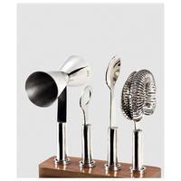 The Mary Jurek Cosmo Bar 4 pc Set On Wood Base features a jigger, bottle opener, spoon, and strainer standing upright in a sleek wooden holder against a plain background.