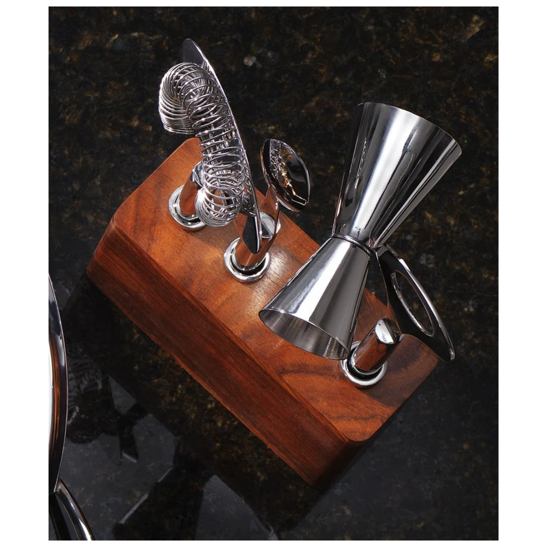 The Mary Jurek Cosmo Bar 4 pc Set On Wood Base features stainless steel bar tools—a cocktail jigger, spiral strainer, and stirring spoon—beautifully arranged on a wooden stand atop a dark, reflective countertop.