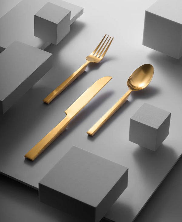 The Ricci Hover Flatware 5 Piece Setting in matte gold—a fork, knife, spoon, salad fork, and teaspoon—rests on a gray surface, accented by white geometric cubes for a sleek, modern touch.