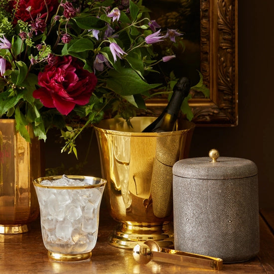 A gold-rimmed glass with ice, the Aerin Shagreen Ice Bucket, embossed shagreen barware, and a floral arrangement with red and purple flowers bring luxury to a home bar vignette on a wooden table.