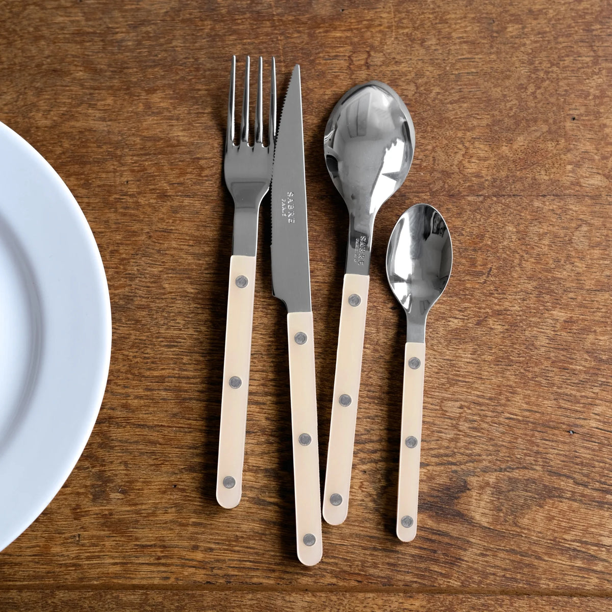 The Sabre Bistrot Pearly 5 Piece Setting - Shiny, featuring beige handles and silver rivets, is arranged on a wooden table beside a white plate.