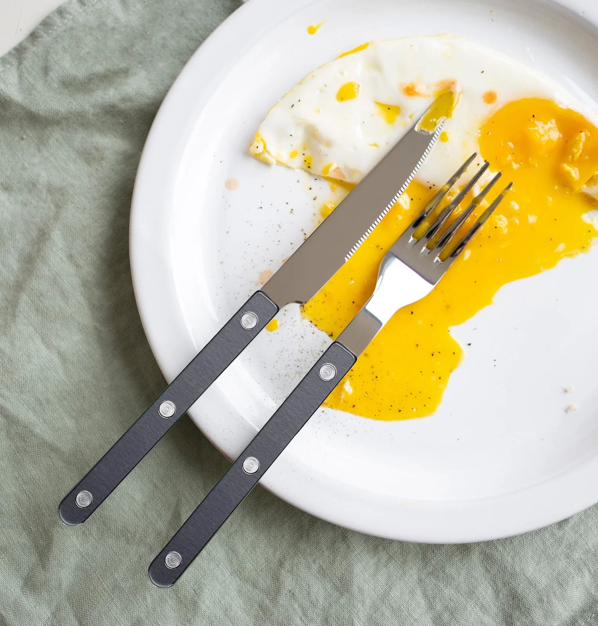 A partially eaten fried egg with runny yolk rests on a white plate, accompanied by a Sabre Bistrot Pearly 5 Piece Setting - Shiny cutlery set. The plate is placed on a light green fabric surface.
