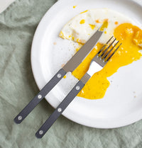 A partially eaten fried egg with runny yolk rests on a white plate, accompanied by a Sabre Bistrot Pearly 5 Piece Setting - Shiny cutlery set. The plate is placed on a light green fabric surface.