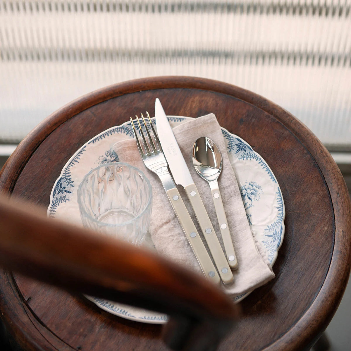A Bistrot Solid Flatware 5 Piece Setting - Shiny by Sabre is arranged with a glass and patterned plate on a beige napkin, displayed on a round wooden table near a sunlit window with blinds.