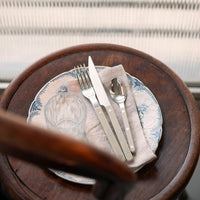 A Bistrot Solid Flatware 5 Piece Setting - Shiny by Sabre is arranged with a glass and patterned plate on a beige napkin, displayed on a round wooden table near a sunlit window with blinds.