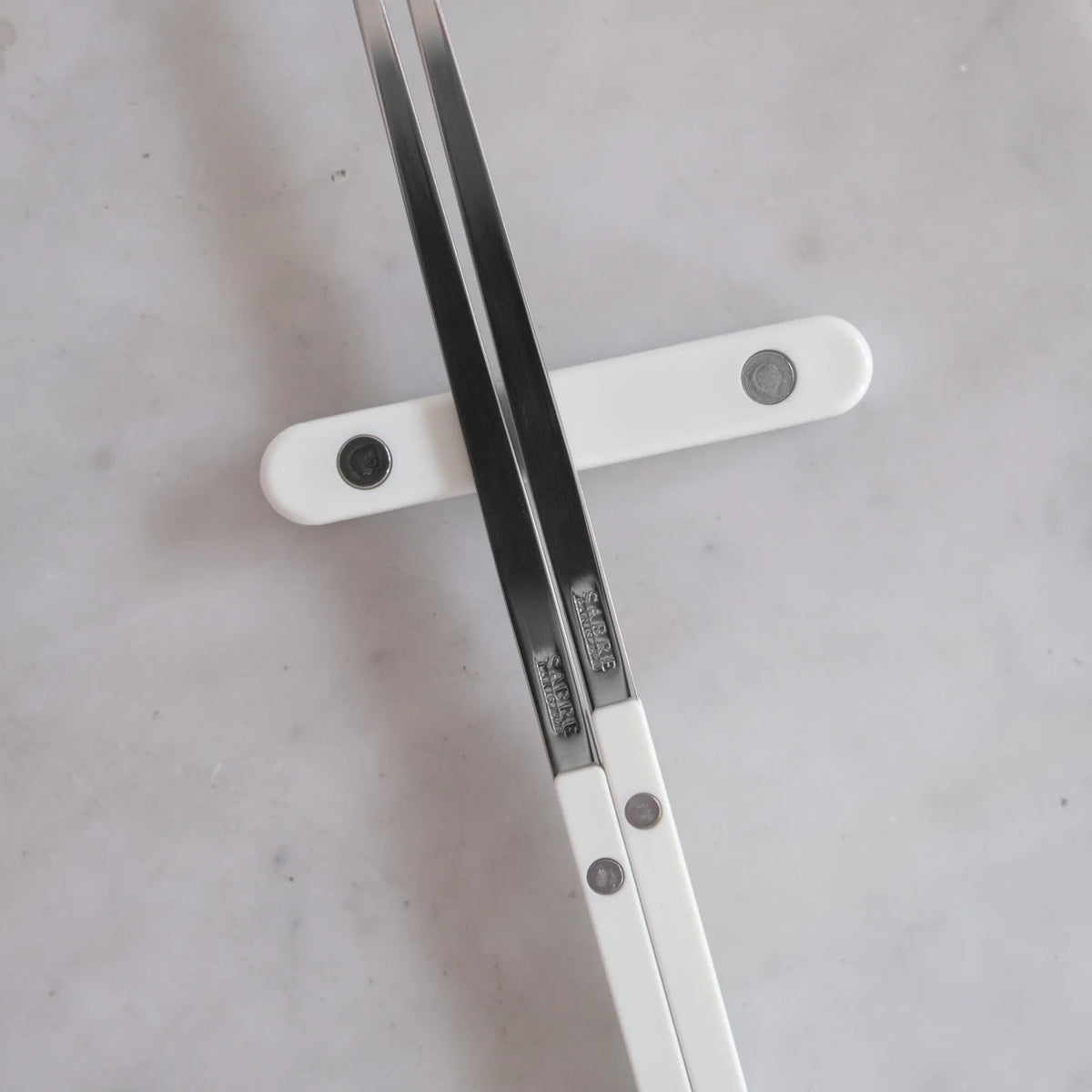 Two Bistrot Solid Chopsticks by Sabre with white handles rest on a white chopstick holder atop a light marble surface, illustrating the refined elegance and the fusion of French craftsmanship with classic Asian tableware.