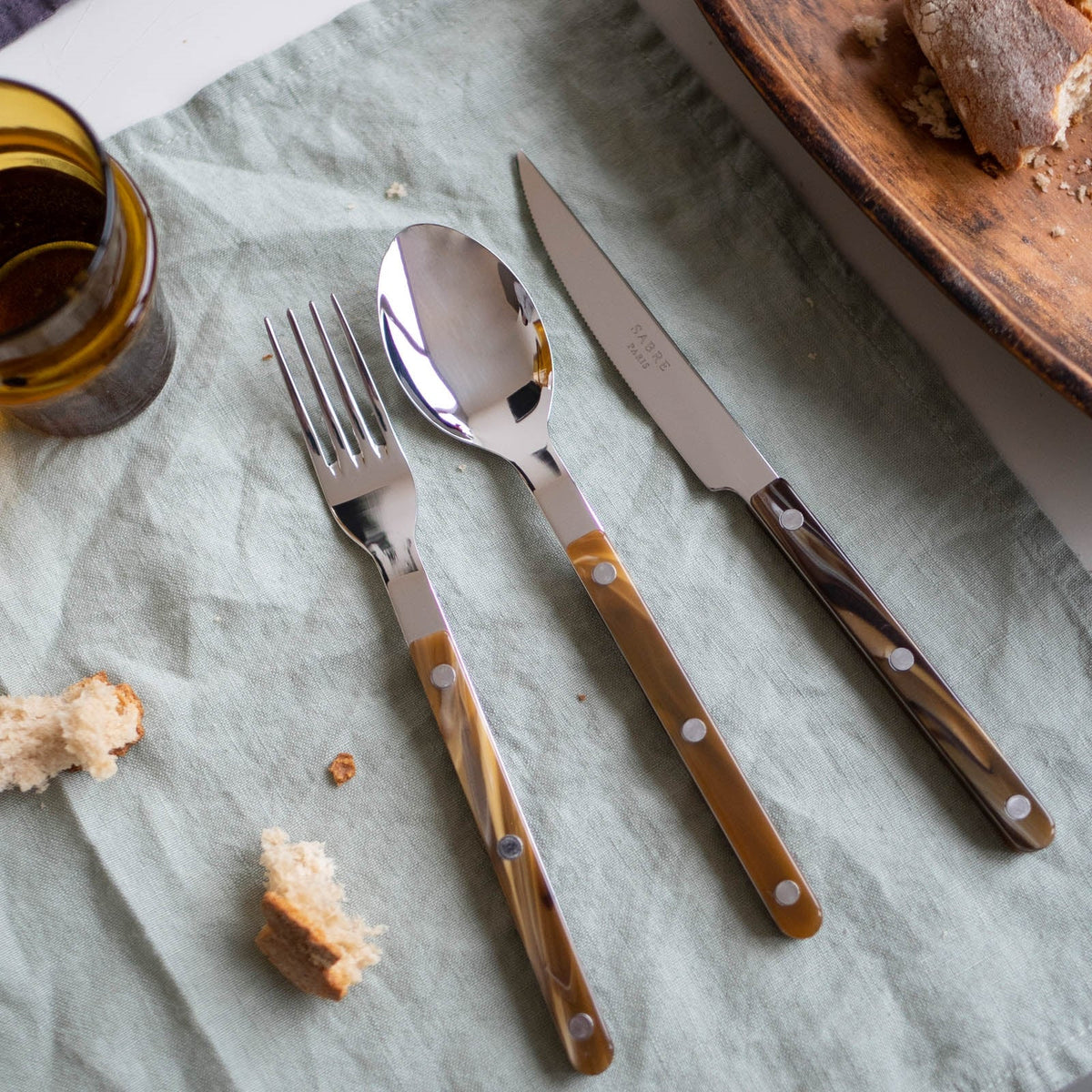 A Sabre Bistrot Flatware 5 Piece Set - Buffalo with brown handles is arranged on a light green cloth, with bread crumbs, a piece of bread, and part of a glass nearby.
