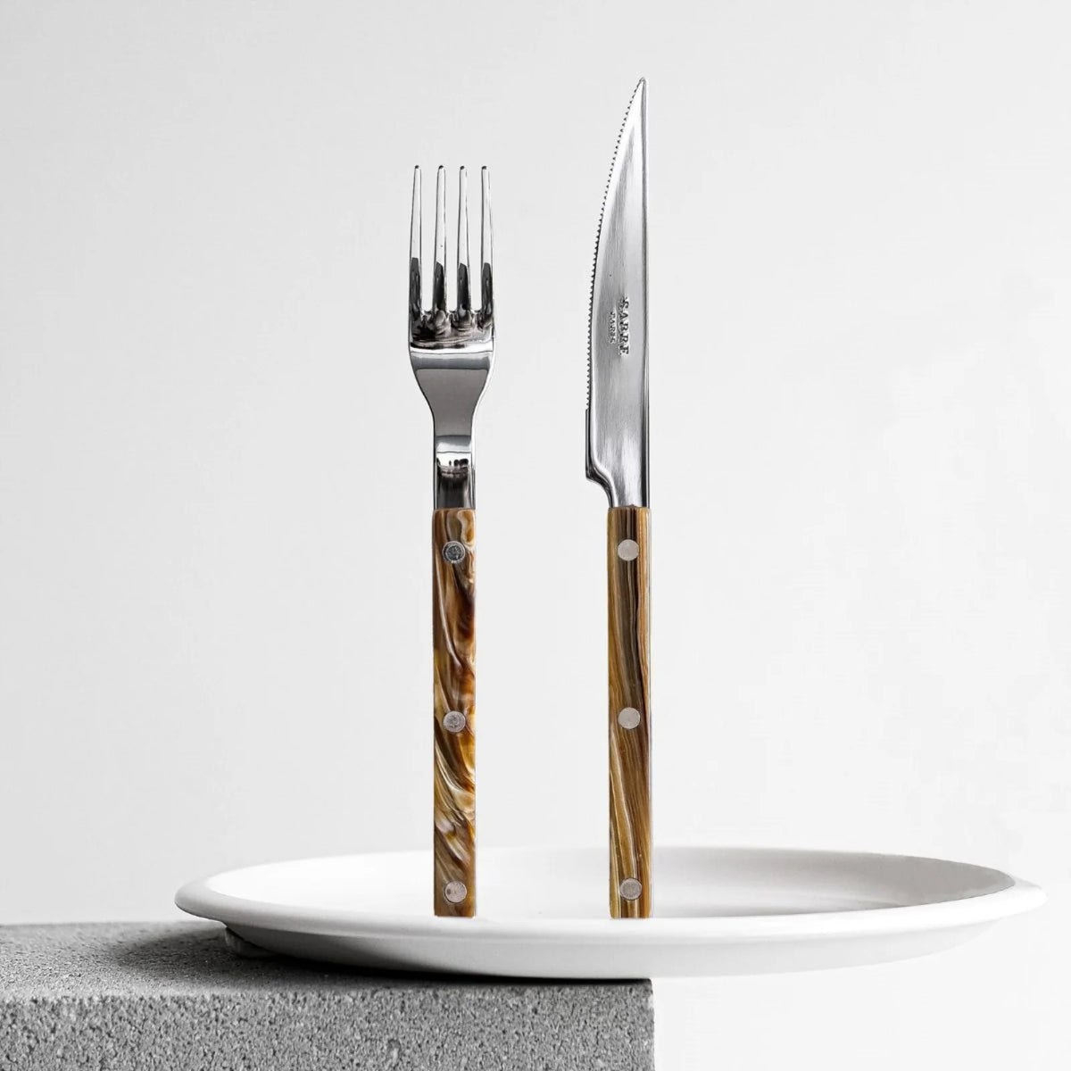The Sabre Bistrot Flatware 5 Piece Set – Buffalo, with brown marbled handles, stands upright on a white plate atop a gray concrete surface against a plain white background.