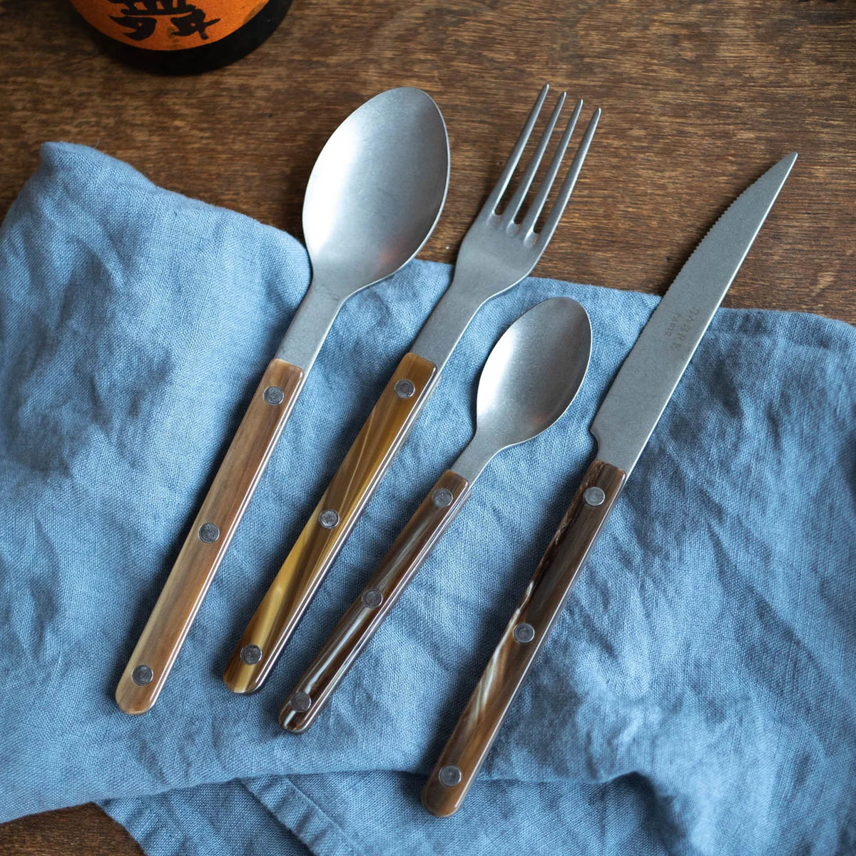 A Sabre Bistrot Flatware 5 Piece Set - Buffalo by Sabre, featuring brown wooden handles, is neatly arranged on a wrinkled blue cloth napkin on a wooden surface. A partial bottle appears in the top left corner.