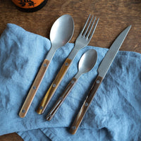 A Sabre Bistrot Flatware 5 Piece Set - Buffalo by Sabre, featuring brown wooden handles, is neatly arranged on a wrinkled blue cloth napkin on a wooden surface. A partial bottle appears in the top left corner.