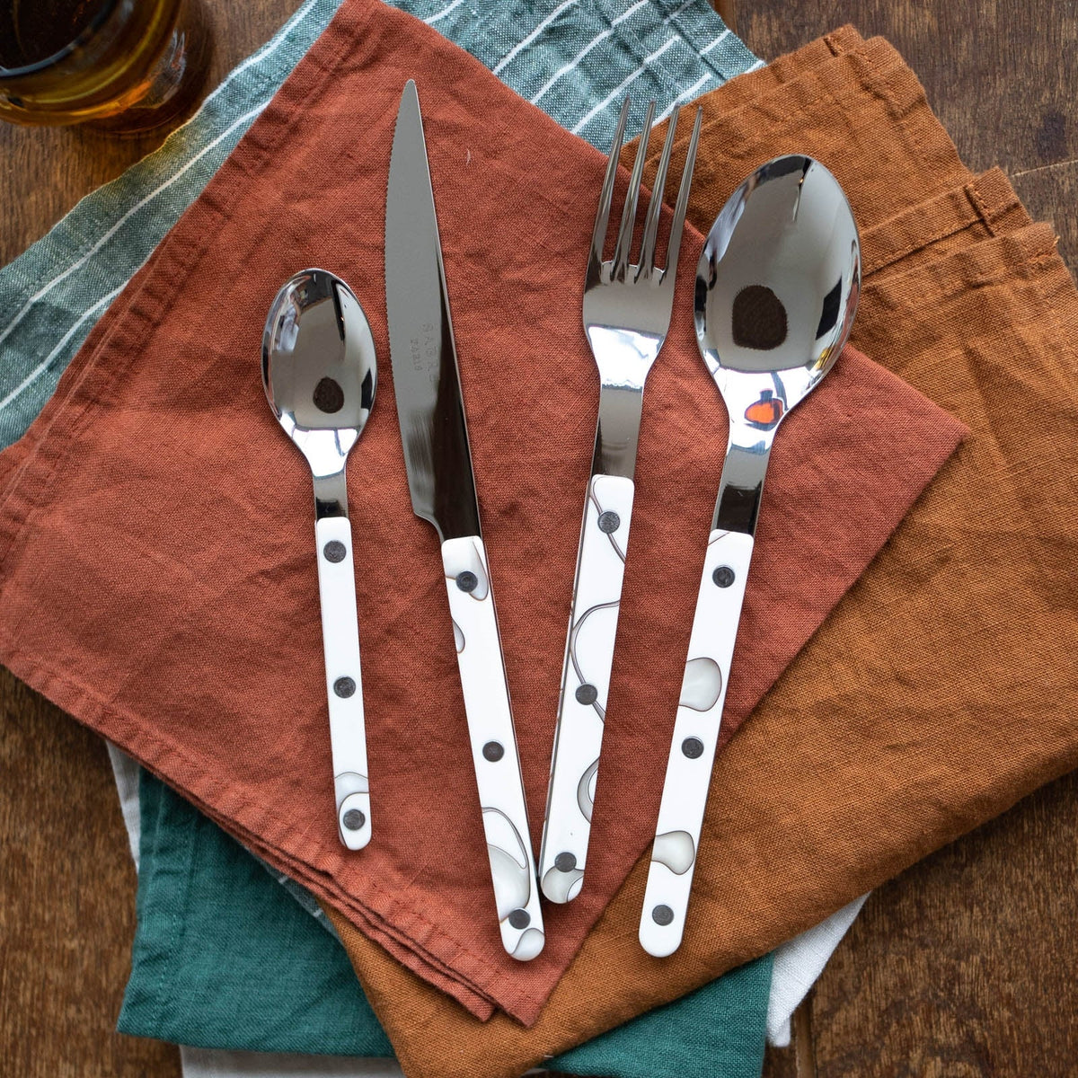 The Sabre Bistro Dune Flatware 5-Piece Setting rests on folded orange, brown, and green cloth napkins atop a wooden surface.