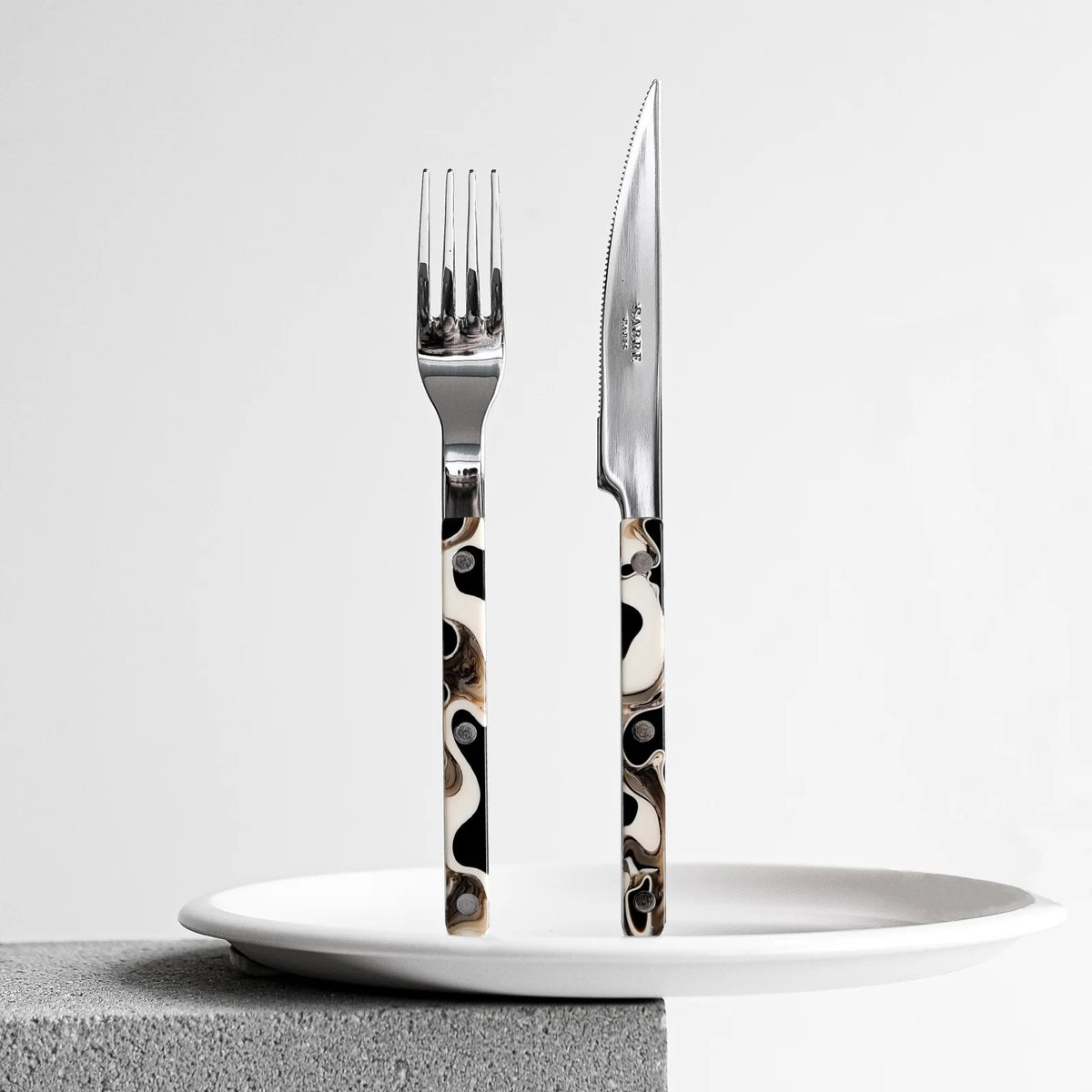 The Sabre Bistro Dune Flatware 5 Piece Setting, featuring marbled black, white, and brown handles, stands upright on a white plate atop a gray textured surface against a plain background.
