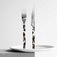 The Sabre Bistro Dune Flatware 5 Piece Setting, featuring marbled black, white, and brown handles, stands upright on a white plate atop a gray textured surface against a plain background.