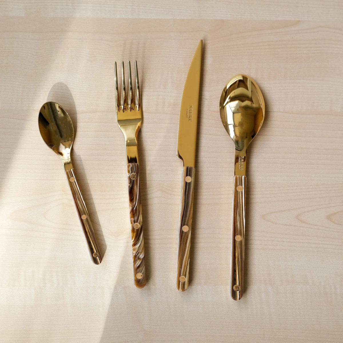 The Sabre Bistrot Gold Flatware 5 Piece Setting – Buffalo features a small spoon, fork, knife, large spoon, and dessert fork with elegant luxury acrylic handles arranged on a light wooden surface.
