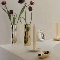 Two tall vases with circular cutouts, one gold and one silver, hold dark purple tulips on a white surface. Two gold candle holders with ivory candles and the Greg Natale Pietra Catchall complete the organic design arrangement.