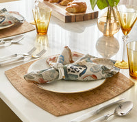 A close-up of an elegant table setting featuring the Kim Seybert Giverny Napkin Set of 4 - Multi, styled with a white plate, gold utensils, amber glasses, and croissants on a wooden board in the background.