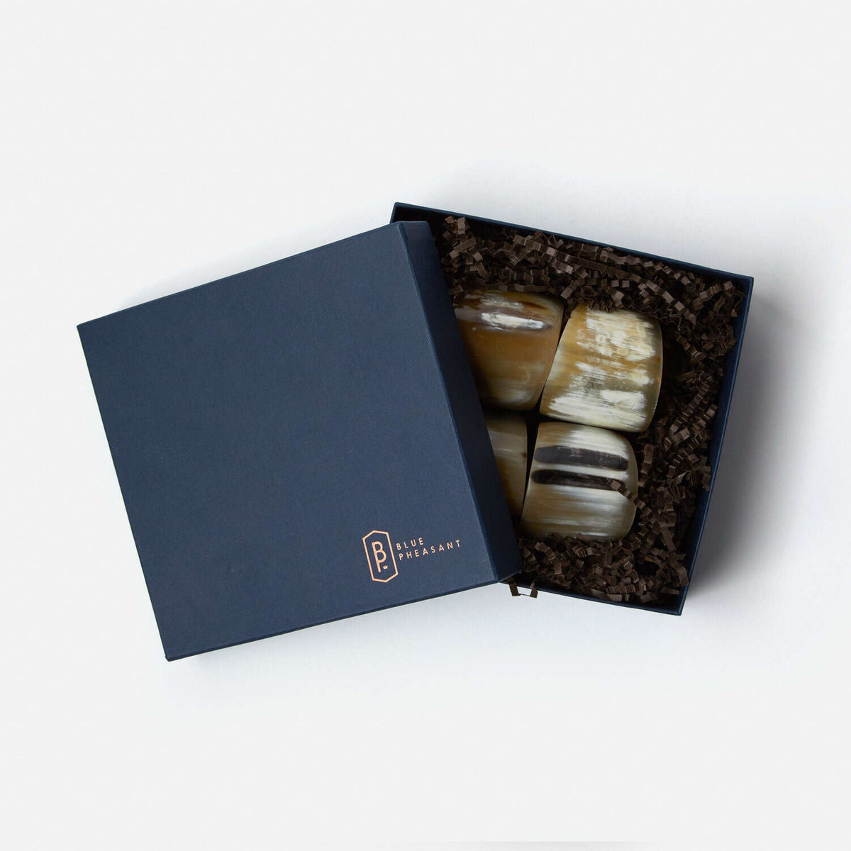A navy blue Blue Pheasant box, partially open on a white background, holds four polished, handmade James Natural Horn Napkin Rings nestled in brown crinkle paper.