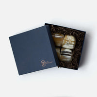 A navy blue Blue Pheasant box, partially open on a white background, holds four polished, handmade James Natural Horn Napkin Rings nestled in brown crinkle paper.
