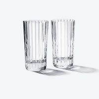 The Baccarat Harmonie Barware Set of 2 features tall, clear highball glasses with vertical faceted patterns, standing side by side on a white surface and casting gentle, elongated shadows to the right.