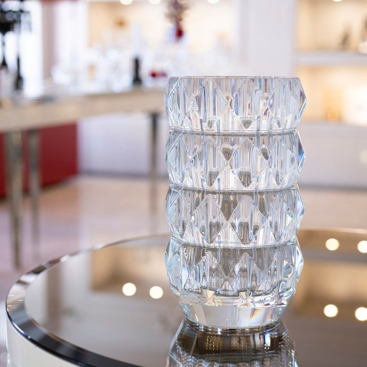 Clear 11″ Louxor Round Vase 230 by Baccarat with a faceted geometric pattern, sitting on a circular reflective table.