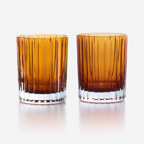 Two orange-colored, faceted Baccarat Harmonie Colors of Joy tumblers from the set of 2 are placed side by side on a reflective white surface against a plain white background.