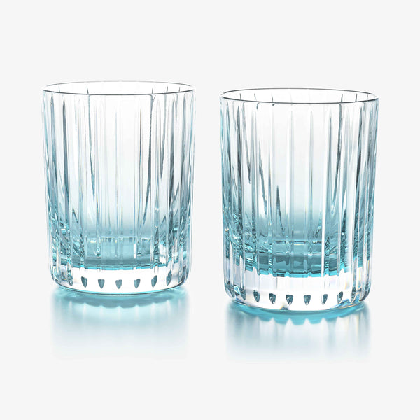 The Harmonie Colors of Joy Set of 2, Turquoise by Baccarat features two clear crystal tumblers with a subtle blue tint and vertical etched lines, elegantly displayed side by side on a white surface—a sophisticated pair perfect for any pastel turquoise-inspired table setting.