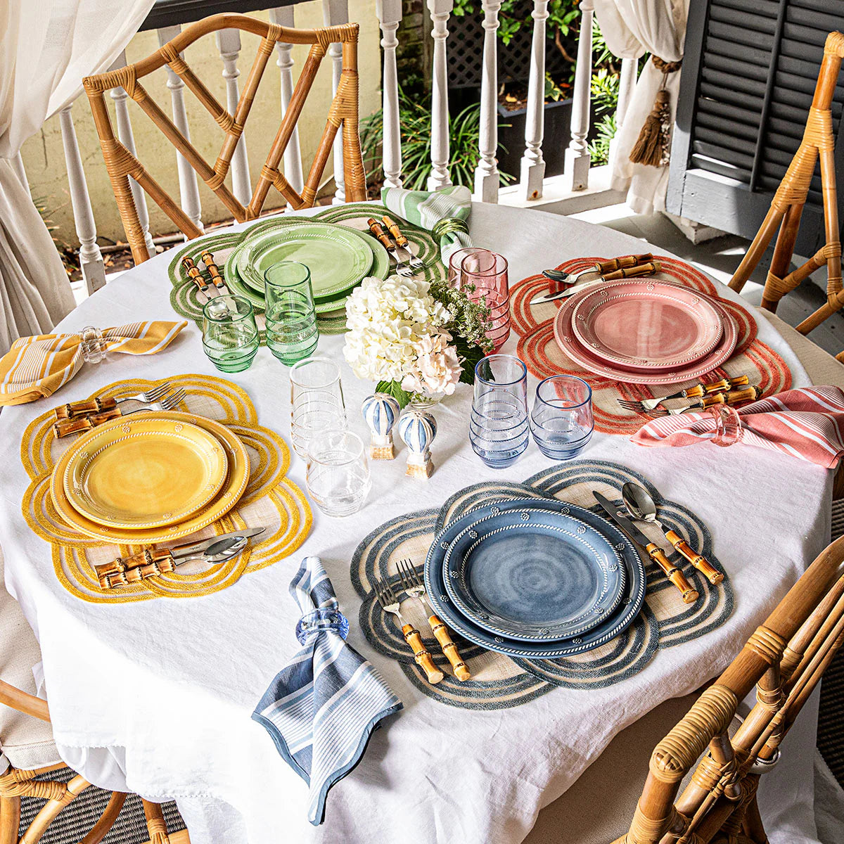 A round table set for four features colorful plates, matching glasses, striped napkins, and Juliska Scallop Stripe Placemats. A white tablecloth, center flowers, and bamboo chairs complete this bright outdoor scene with a touch of quatrefoil style.