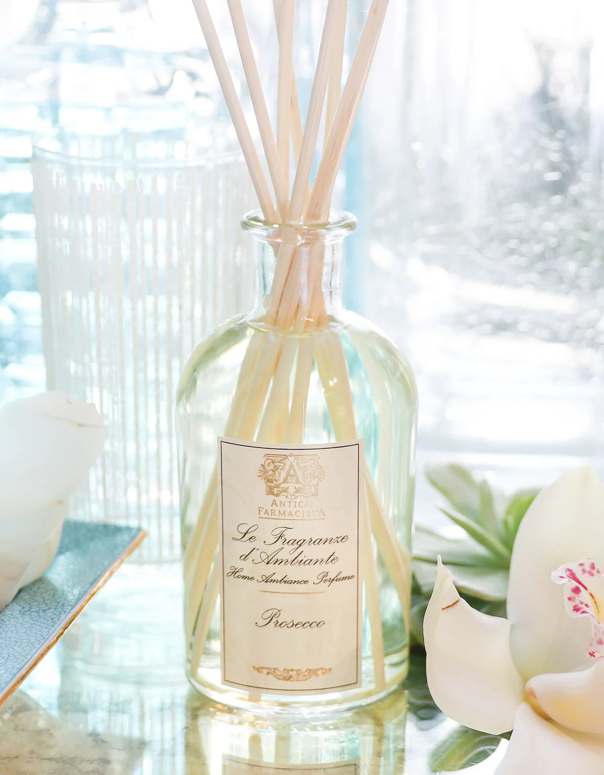 A glass bottle of Antica Farmacista Prosecco Diffuser by Harper stands with thin reeds on a surface, accompanied by white flowers and blurred glass decor, creating an elegant home fragrance display in the background.
