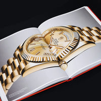 An open copy of "The Book of Rolex" by ACC ART BOOKS / SIMON & SCHUSTER shows a detailed photo of a gold Rolex watch with fluted bezel, gold bracelet, and day-date display, set against a plain gray background.