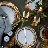 A beautifully set table with L'Objet Corde Dinnerware Gold, gold-rimmed chargers, grey napkins in rings, candles, and floral accents on a dark green cloth highlights the collection’s elegance and modern style.