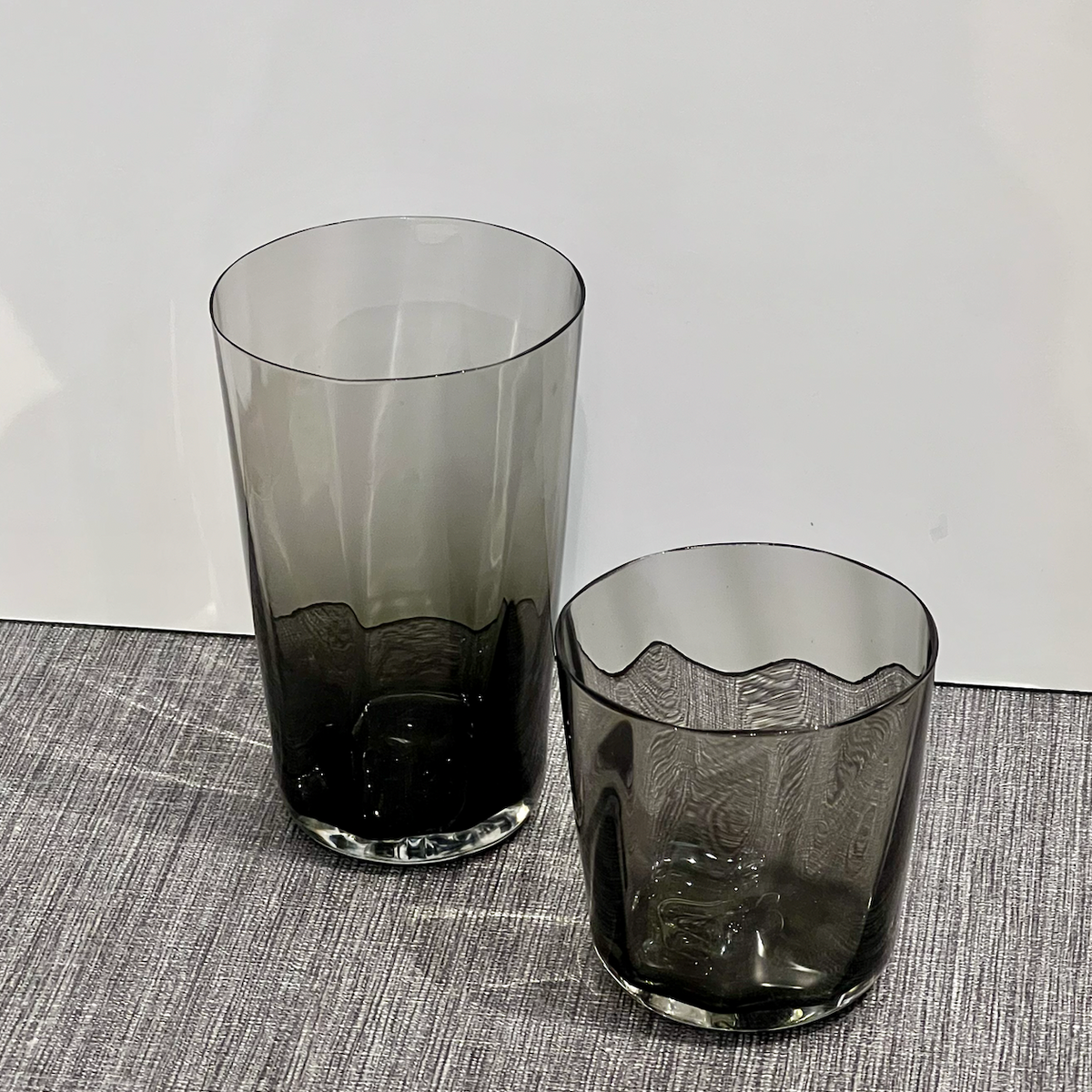 Two Optic Cups Charcoal by Gary Bodker—one tall and cylindrical, the other shorter with a wavy rim—sit on a textured grey surface against a plain white background.