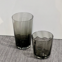 Two Optic Cups Charcoal by Gary Bodker—one tall and cylindrical, the other shorter with a wavy rim—sit on a textured grey surface against a plain white background.