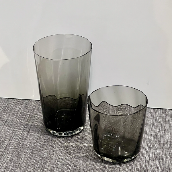 Two Optic Cups Charcoal by Gary Bodker—one tall and cylindrical, the other shorter with a wavy rim—sit on a textured grey surface against a plain white background.