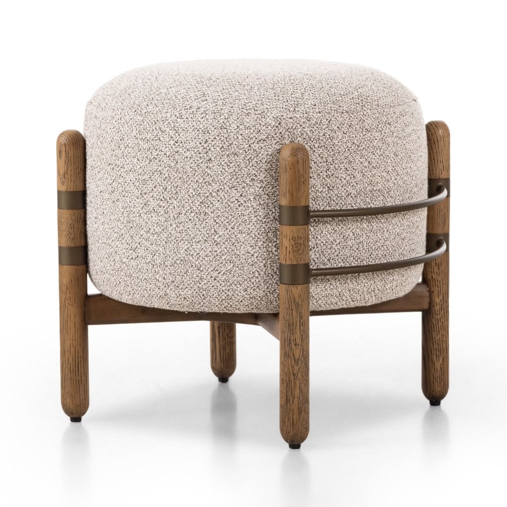 The Benson Ottoman 21″ by Four Hands features a round silhouette upholstered in light beige Astor Stone fabric with a textured finish, supported by four wooden legs and accented with two horizontal distressed natural bronze bands around the base.