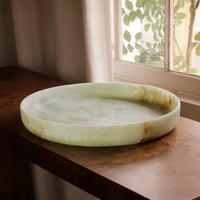 The Beatriz Ball Resin Giada Round Tray in Jade Swirl, with natural brown accents, sits on a wooden table near a window with white curtains and leafy plants outside.