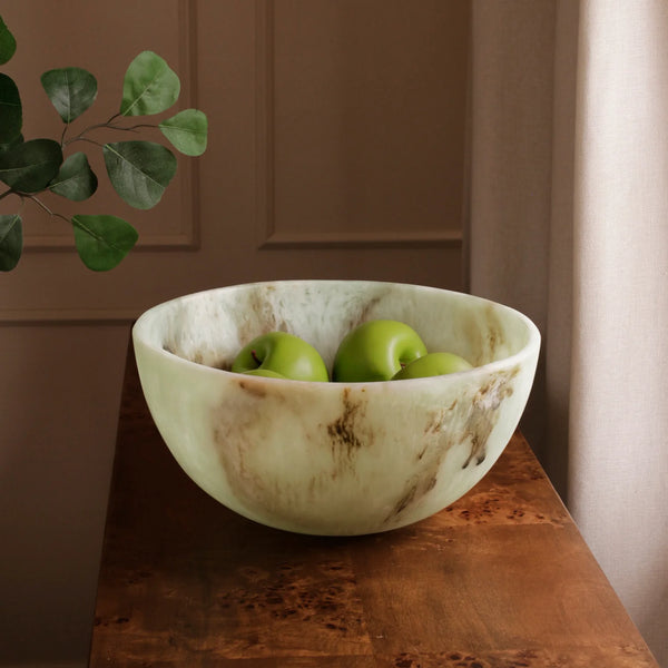 The Beatriz Ball Resin Giada Bowl Jade Swirl - Large sits on a wooden surface, filled with three green apples. A leafy plant appears on the left, and sunlight streams through a window with beige curtains.