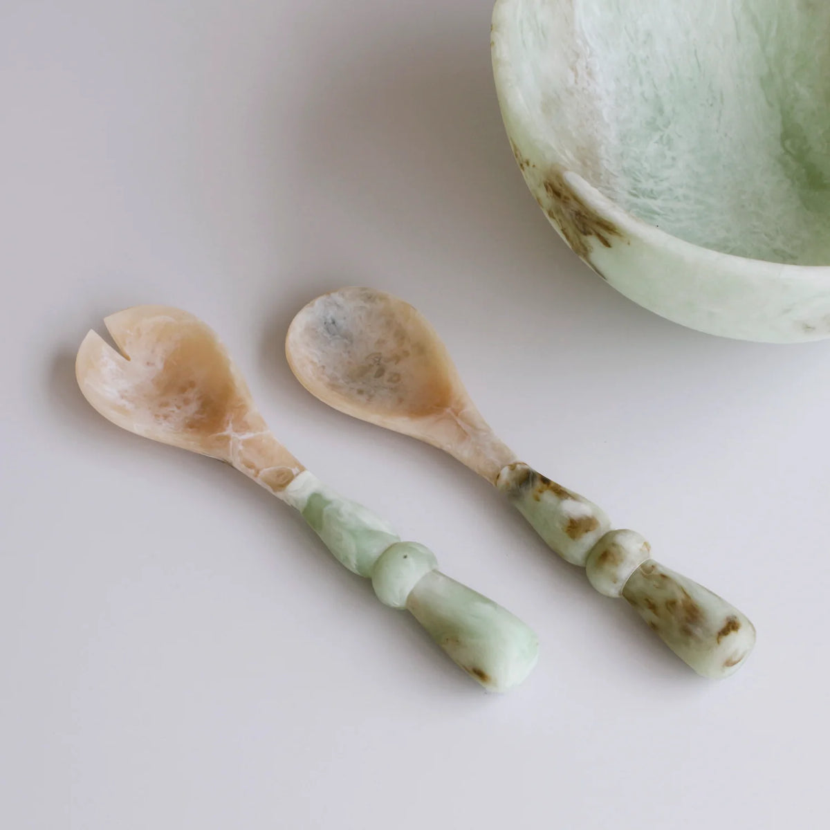 Two Beatriz Ball Resin Giada Salad Server Sets in Jade Swirl, featuring elegantly curved handles, are placed beside a coordinating bowl on a smooth white surface, highlighting the refined beauty of jade-inspired tableware.