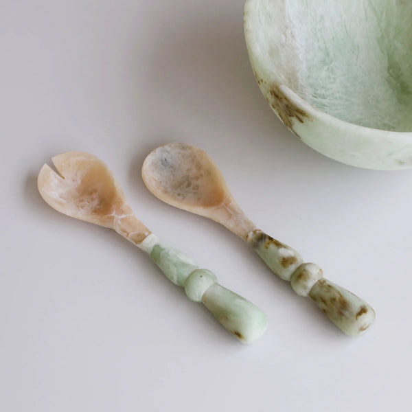 Two Beatriz Ball Resin Giada Salad Server Sets in Jade Swirl, featuring elegantly curved handles, are placed beside a coordinating bowl on a smooth white surface, highlighting the refined beauty of jade-inspired tableware.