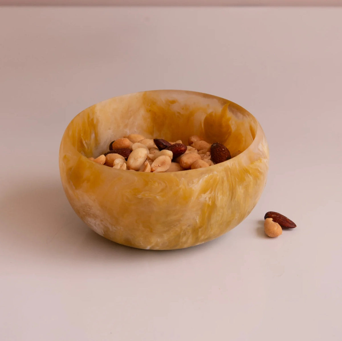 The Beatriz Ball Resin Sorbet Mini Bowl - Matcha, marbled in yellow, is filled with mixed nuts and placed on a pale surface with scattered nuts—perfect for highlighting distinctive tableware.