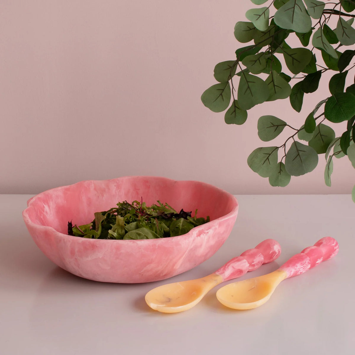 A pink marbled salad bowl with leafy greens sits on a white surface beside the Beatriz Ball Resin Sorbet Salad Server Set - Raspberry, as eucalyptus branches hang above against a pale wall.