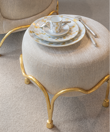 A stack of elegant Marie Daâge Zephyr 1 Dinnerware by Marie Daâge, along with a cup and silverware, sits atop a beige ottoman with gold legs on a light carpeted floor.