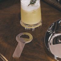 A yellow cocktail with ice and a sprig garnish rests on a wooden table beside the Oh De Stainless Steel Bottle Opener by Christofle, a round metal coaster nearby—an elegant addition to any home bar setup.
