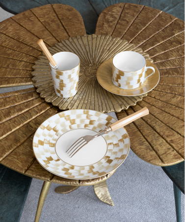 The Marie Daâge Zephyr 1 Dinnerware set by Marie Daâge features a gold and white checkered hand-painted porcelain plate, cup, and saucer, elegantly accompanied by wooden-handled cutlery on a gold flower-shaped table. Made in France, this sophisticated ensemble adds refined charm to any dining setting.