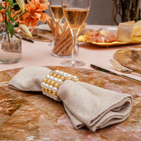 A beige napkin with the Pearl Cuff Napkin Ring Gold Set of 4 by Von Gern Home rests on a marbled placemat, complemented by champagne glasses, breadsticks, flowers, and a platter of bread and cured meats for an elegant table setting.