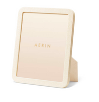 The Aerin Modern Shagreen Frame Cream is a rectangular tabletop frame with an embossed shagreen texture in beige, featuring a stand and displaying a card printed with 'AERIN' in gold at the center for minimalist appeal.