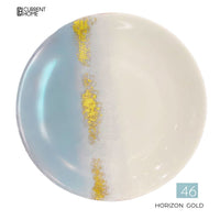 The Marie Daâge Horizon Gold Dinnerware features a round porcelain plate with a light blue-to-white gradient, a vertical gold and white speckled band, and the text "CURRENT HOME" and "46 HORIZON GOLD.
