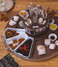 A Christofle MOOD Asia Tray - Stainless Steel holds assorted Asian dishes—dumplings, sushi, vegetables, and jelly desserts—with a central holder for forks and spoons. The platter is placed on a wooden table.