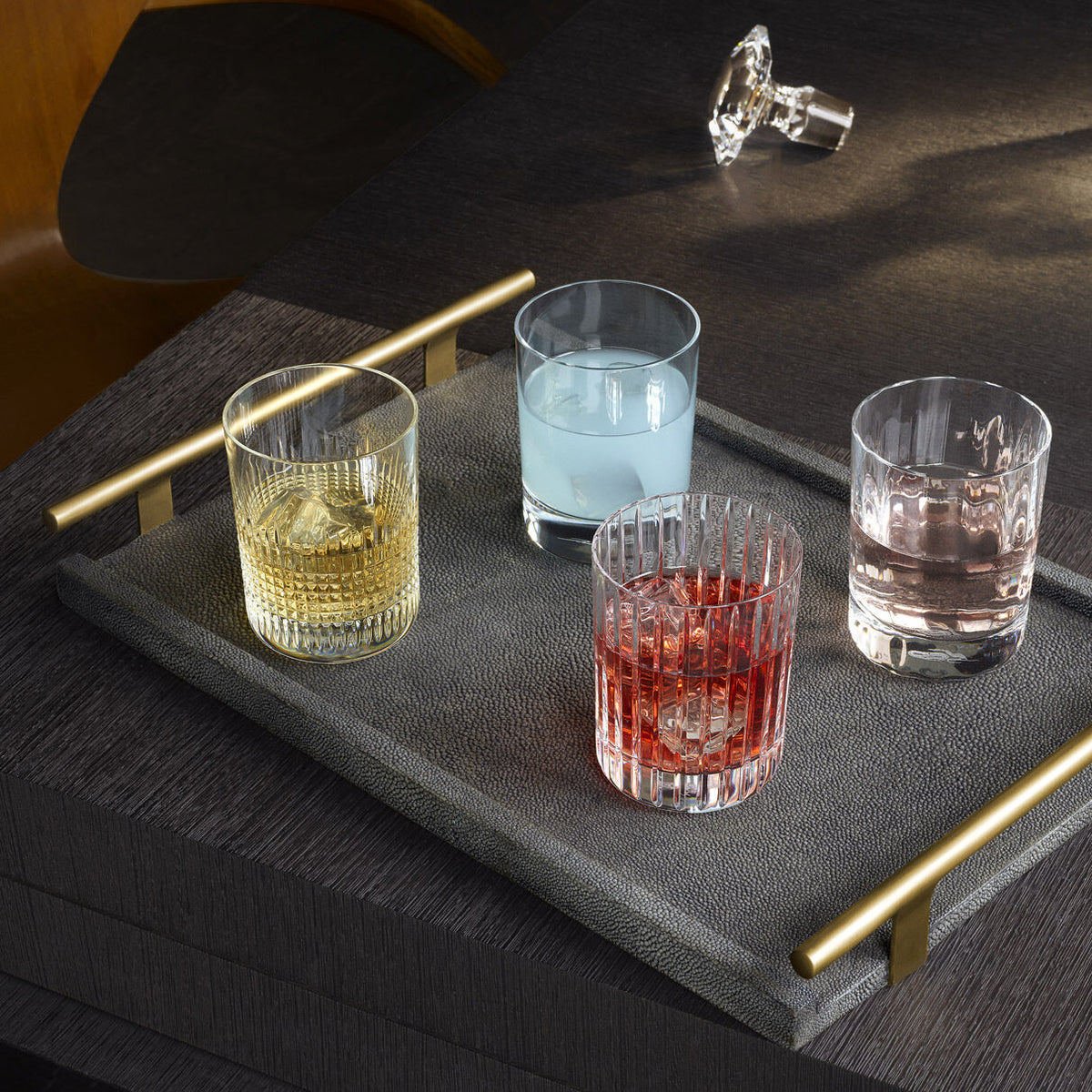 A gray tray with gold handles displays the Baccarat 4 Elements DOF Gift Set—four crystal Double Old Fashioned tumblers, each filled with a different colored drink—resting on a dark wooden table with a chair and glass stopper behind.