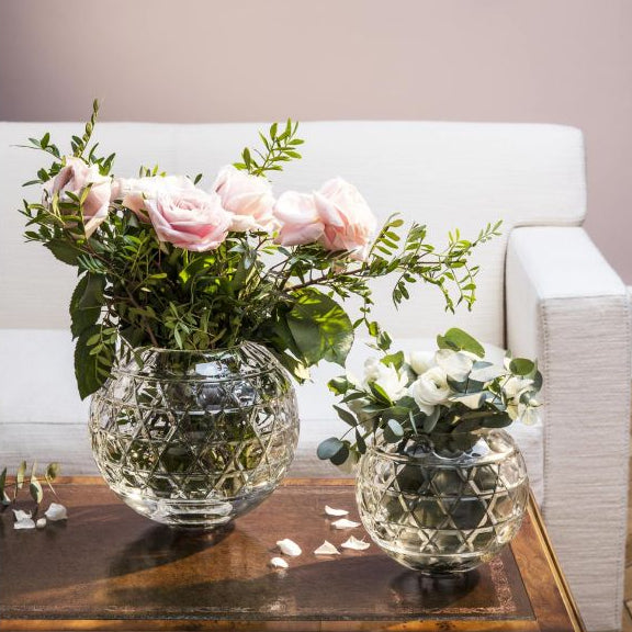 Two round, clear glass Royal Vase - Saint-Louis/Hermes vases with a geometric cut pattern, each holding a floral arrangement, placed on a wooden table.