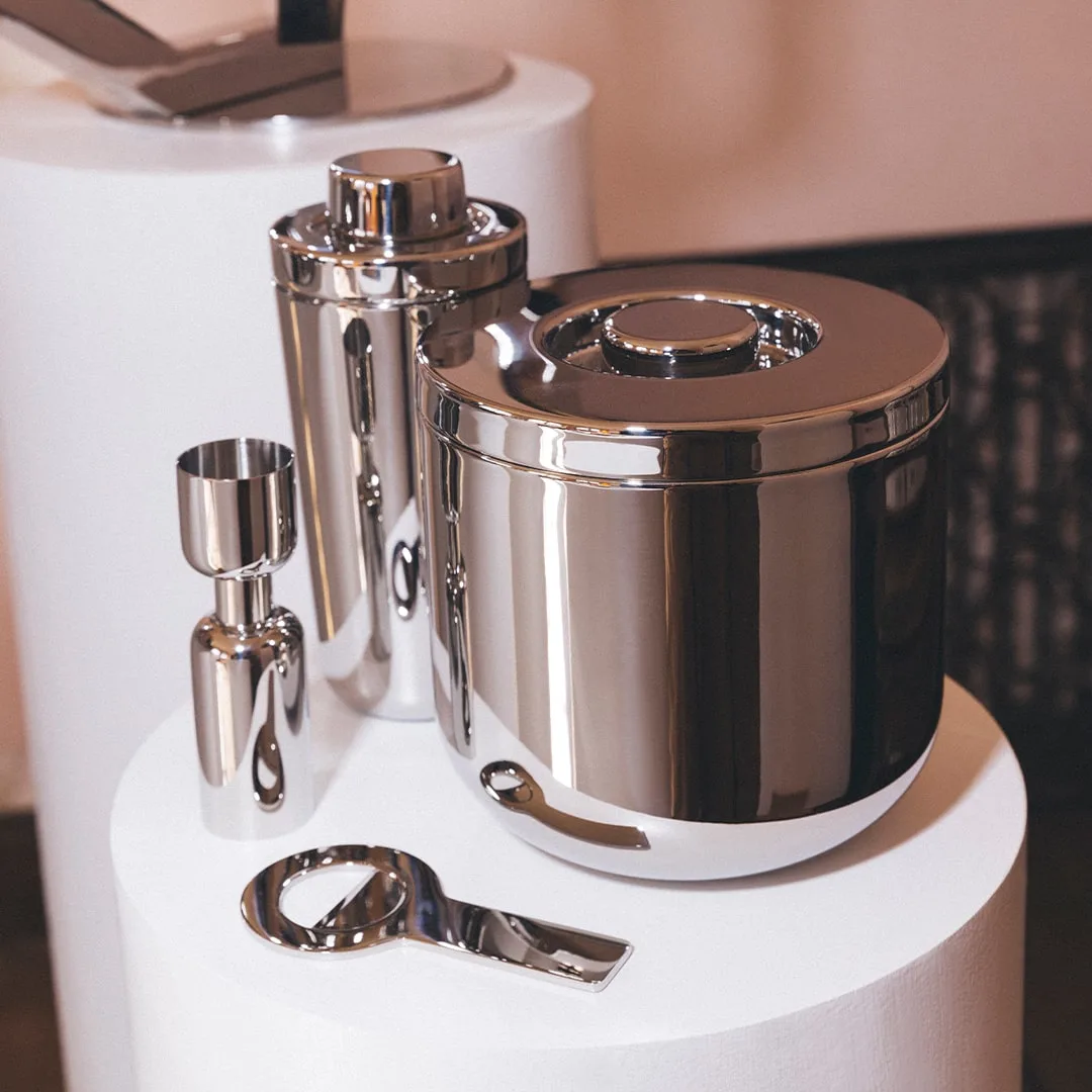Christofle’s Oh De Stainless Steel Bottle Opener, shown with a matching ice bucket, cocktail shaker, and jigger on a white pedestal, is perfect for elevating any home bar against a neutral backdrop.